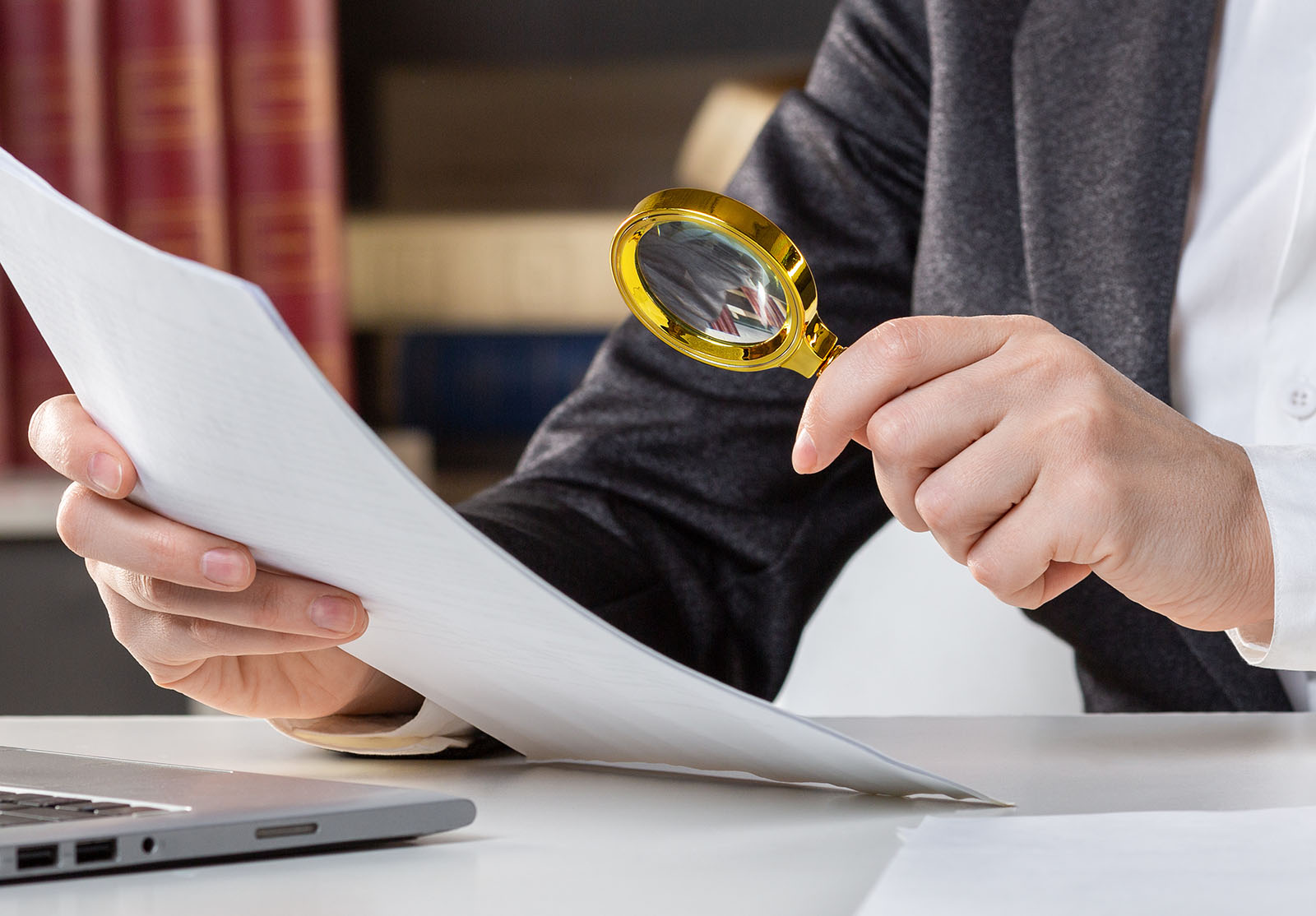 Main féminine avec une loupe dorée et un papier sur le bureau à côté d'un ordinateur portable ouvert, gros plan, mise au point sélectionnée. Loupe, contrat, avocat, attention, audit, vigilance, assureur, concept de paperasse.