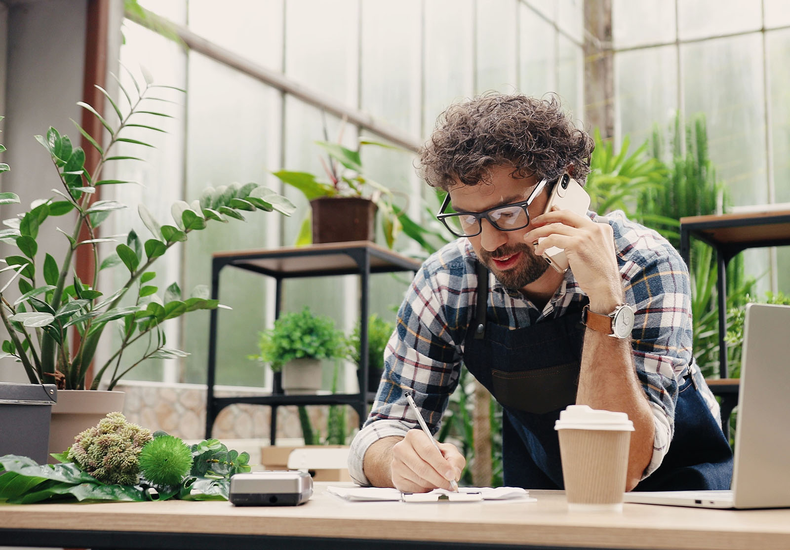 Un homme d'affaires caucasien heureux parle au téléphone portable tout en se tenant debout en tablier dans un petit centre floral et en notant les détails de la commande. Fleuriste joyeux appelant sur son smartphone au travail. Concept d'entreprise personnelle